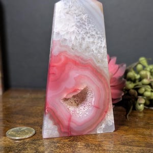 May include: A polished agate obelisk with a flat top. The stone features concentric bands of red, white, and gray, with a central geode cavity. The obelisk is displayed on a wooden surface, with a coin visible.