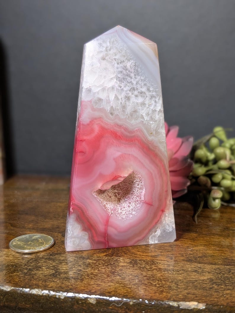 May include: A polished agate obelisk with a flat top. The stone features concentric bands of red, white, and gray, with a central geode cavity. The obelisk is displayed on a wooden surface, with a coin visible.