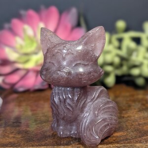 May include: A carved, lavender-colored cat figurine. The cat is sitting upright with its tail curled. The figurine has pointed ears and a smiling face. The cat is made of a translucent material, and the details are well-defined.