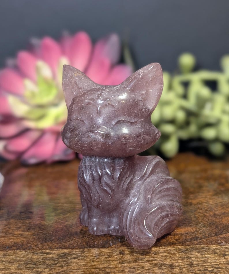 May include: A carved, lavender-colored cat figurine. The cat is sitting upright with its tail curled. The figurine has pointed ears and a smiling face. The cat is made of a translucent material, and the details are well-defined.
