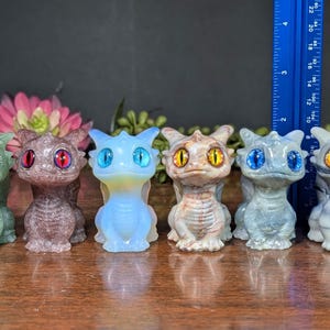 Pode incluir: Seis pequenas figuras de drag&otilde;es coloridos, verdes, roxos, azuis, bege e cinzentos. Cada drag&atilde;o tem olhos grandes e detalhados de cores diferentes. Uma r&eacute;gua azul &eacute; vis&iacute;vel no fundo. As figuras t&ecirc;m cerca de 10 cm de altura.