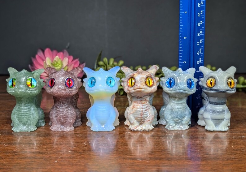 Pode incluir: Seis pequenas figuras de drag&otilde;es coloridos, verdes, roxos, azuis, bege e cinzentos. Cada drag&atilde;o tem olhos grandes e detalhados de cores diferentes. Uma r&eacute;gua azul &eacute; vis&iacute;vel no fundo. As figuras t&ecirc;m cerca de 10 cm de altura.