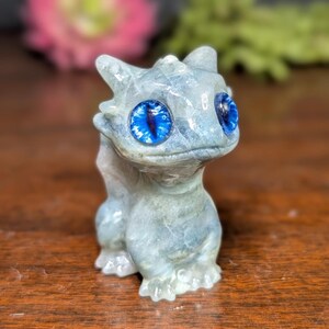 Pode incluir: Estatueta de drag&atilde;o azul claro com olhos grandes e azul brilhante. O drag&atilde;o &eacute; esculpido em um material semelhante a pedra e tem uma superf&iacute;cie detalhada e texturizada. A estatueta est&aacute; em uma superf&iacute;cie de madeira.