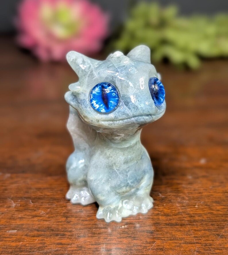 Pode incluir: Estatueta de drag&atilde;o azul claro com olhos grandes e azul brilhante. O drag&atilde;o &eacute; esculpido em um material semelhante a pedra e tem uma superf&iacute;cie detalhada e texturizada. A estatueta est&aacute; em uma superf&iacute;cie de madeira.