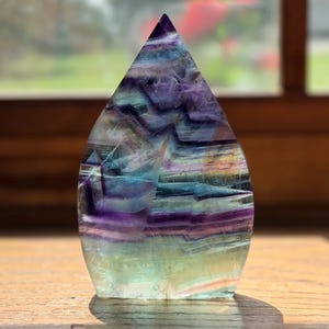 May include: A polished flame-shaped fluorite crystal sculpture featuring distinct horizontal bands of deep purple, vibrant blue, and pale green. The translucent stone displays natural internal fractures and color gradients throughout its structure.
