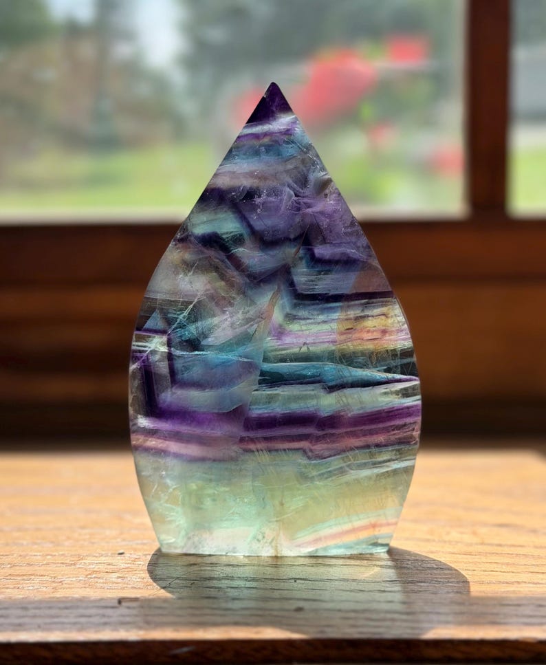 May include: A polished flame-shaped fluorite crystal sculpture featuring distinct horizontal bands of deep purple, vibrant blue, and pale green. The translucent stone displays natural internal fractures and color gradients throughout its structure.