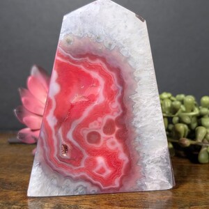 May include: A polished, triangular crystal obelisk with a flat top. The stone features a vibrant red and white banded pattern, with a white crystalline edge. The obelisk is displayed on a wooden surface, with a pink flower and green plant in the background.