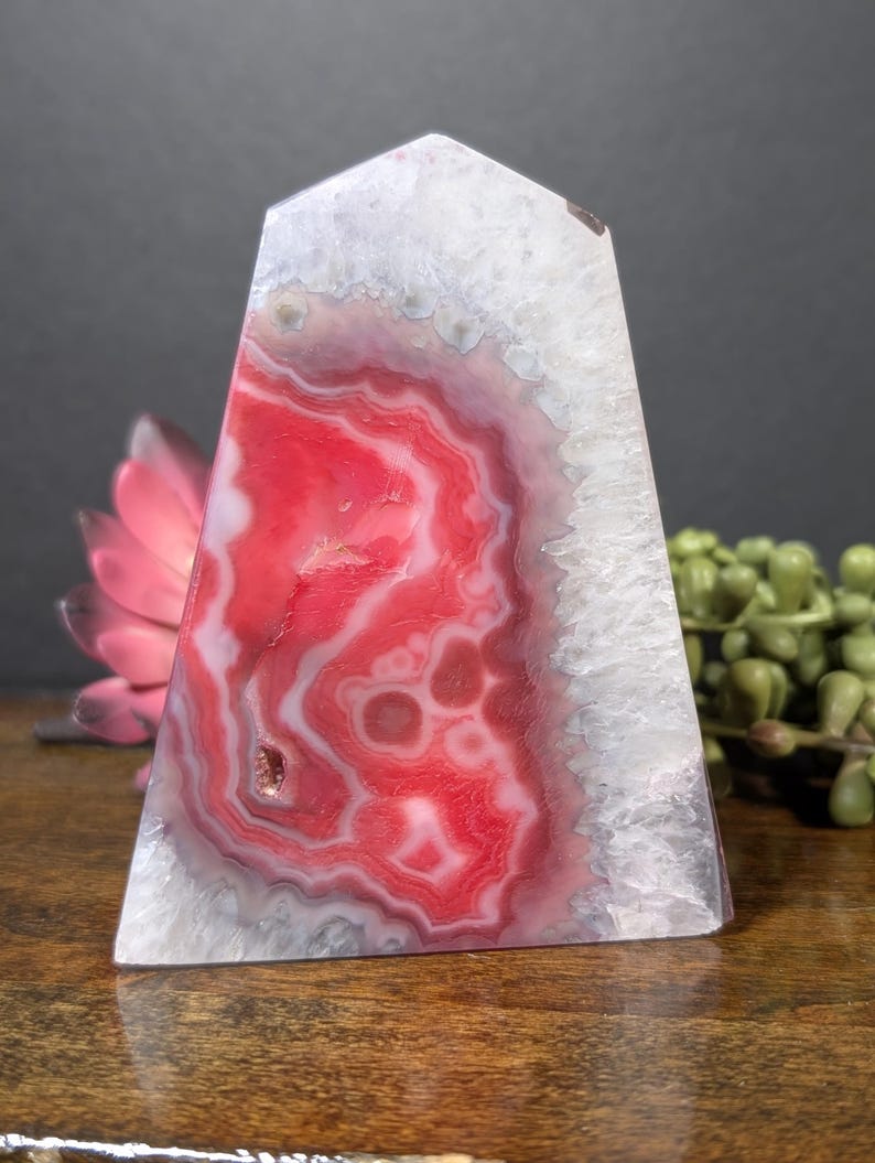 May include: A polished, triangular crystal obelisk with a flat top. The stone features a vibrant red and white banded pattern, with a white crystalline edge. The obelisk is displayed on a wooden surface, with a pink flower and green plant in the background.