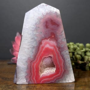 May include: A polished, obelisk-shaped agate stone with a striking red and white pattern. The center features a geode cavity with crystal formations. The stone's colors range from white to deep red, creating a visually appealing display.