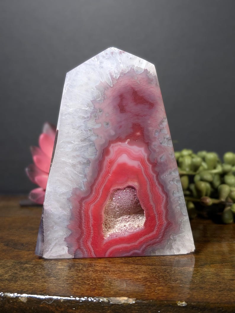 May include: A polished, obelisk-shaped agate stone with a striking red and white pattern. The center features a geode cavity with crystal formations. The stone's colors range from white to deep red, creating a visually appealing display.