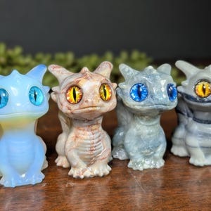 Pode incluir: Uma cole&ccedil;&atilde;o de pequenas figuras de drag&otilde;es detalhadas. Os drag&otilde;es s&atilde;o em v&aacute;rias cores, incluindo azul, cinza e bege, com grandes olhos expressivos em tons de azul e amarelo. Eles t&ecirc;m corpos texturizados e pequenas asas.