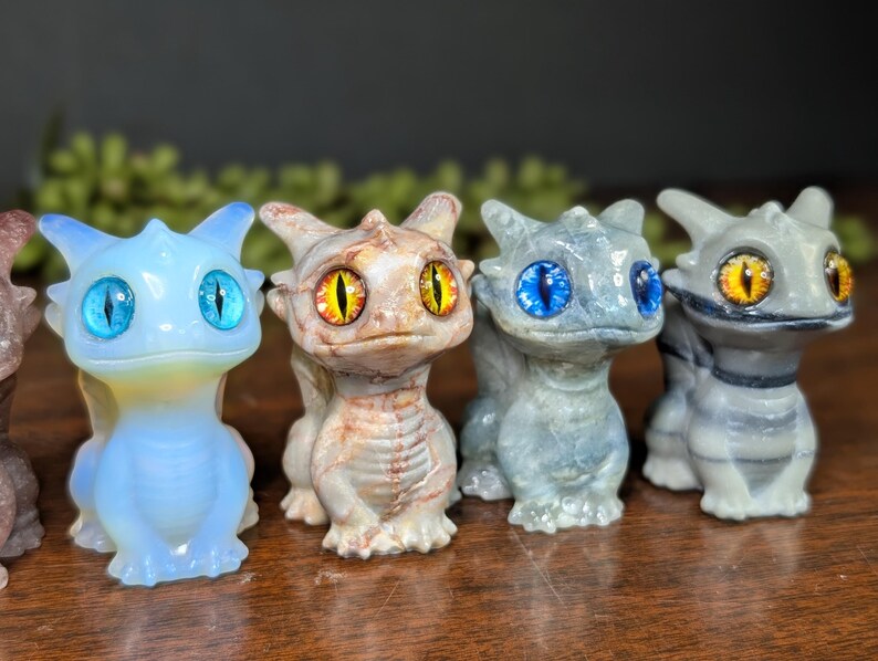 Pode incluir: Uma cole&ccedil;&atilde;o de pequenas figuras de drag&otilde;es detalhadas. Os drag&otilde;es s&atilde;o em v&aacute;rias cores, incluindo azul, cinza e bege, com grandes olhos expressivos em tons de azul e amarelo. Eles t&ecirc;m corpos texturizados e pequenas asas.