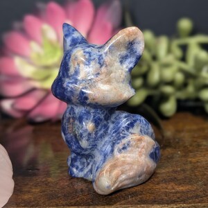 May include: A small, carved fox figurine made of blue sodalite stone with tan and white accents. The fox is sitting upright with its ears perked up, showcasing the natural patterns and colors of the stone. A decorative item.