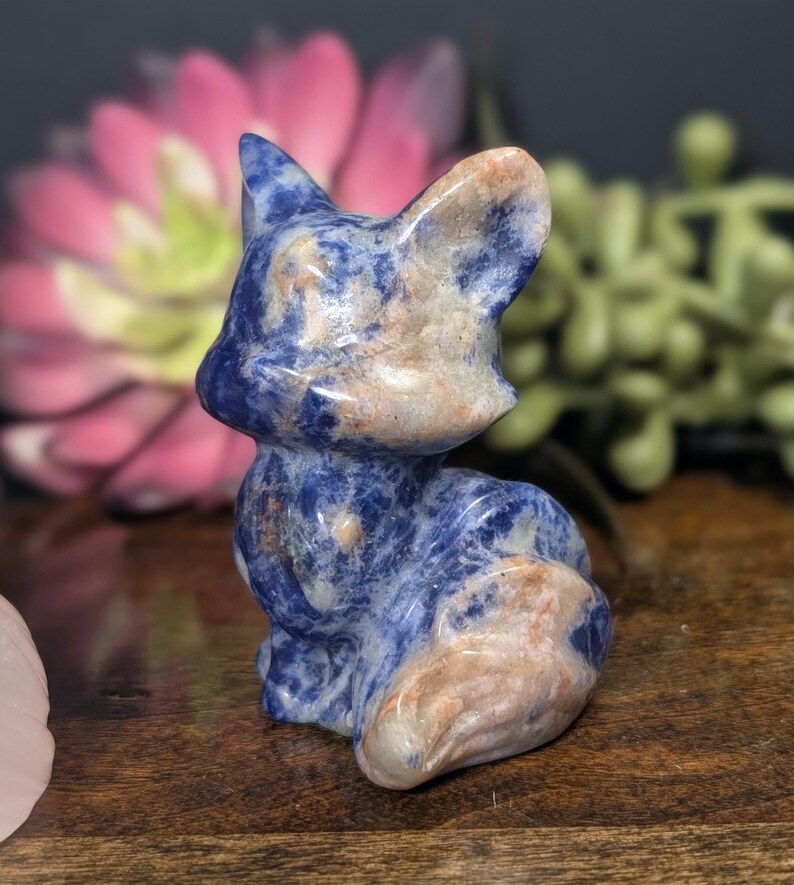May include: A small, carved fox figurine made of blue sodalite stone with tan and white accents. The fox is sitting upright with its ears perked up, showcasing the natural patterns and colors of the stone. A decorative item.