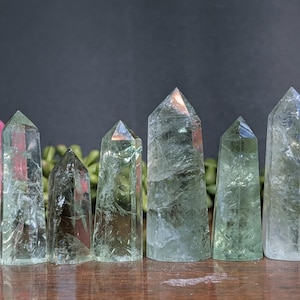 May include: Six green quartz crystal points of varying sizes are displayed, with a blue ruler for scale. The crystals are translucent and have a natural, textured appearance. The tallest crystal is approximately 12.7 cm tall.