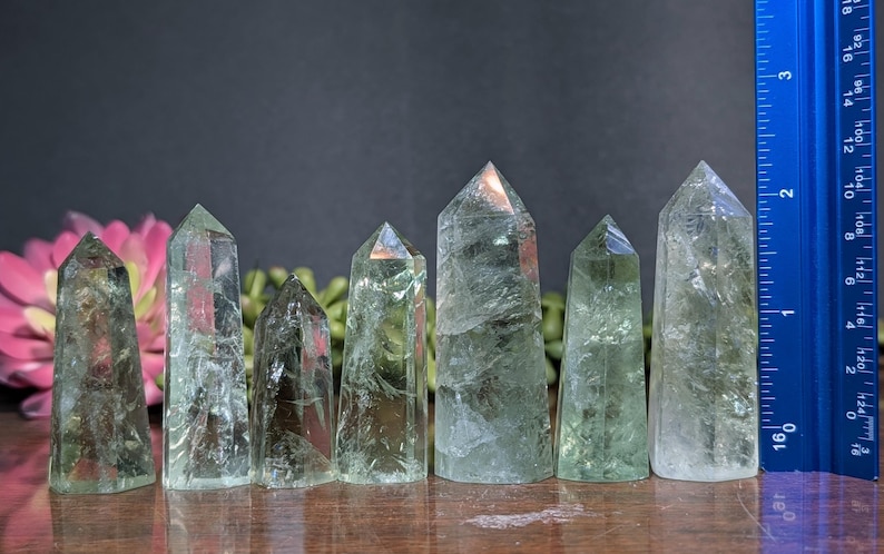 May include: Six green quartz crystal points of varying sizes are displayed, with a blue ruler for scale. The crystals are translucent and have a natural, textured appearance. The tallest crystal is approximately 12.7 cm tall.