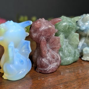 Pode incluir: Uma cole&ccedil;&atilde;o de figuras de drag&otilde;es de cristal esculpidas em v&aacute;rias cores. Os drag&otilde;es t&ecirc;m asas e s&atilde;o em tons de azul, roxo, verde e branco. S&atilde;o exibidos em uma superf&iacute;cie de madeira.