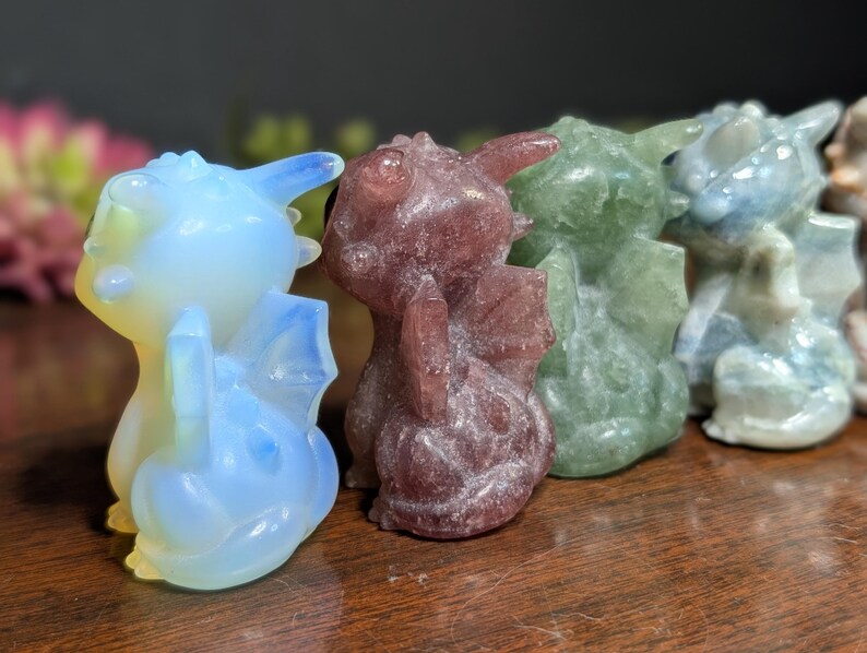 Pode incluir: Uma cole&ccedil;&atilde;o de figuras de drag&otilde;es de cristal esculpidas em v&aacute;rias cores. Os drag&otilde;es t&ecirc;m asas e s&atilde;o em tons de azul, roxo, verde e branco. S&atilde;o exibidos em uma superf&iacute;cie de madeira.