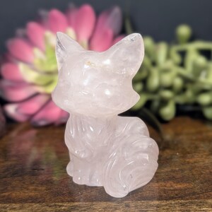 May include: A carved rose quartz fox figurine. The fox is light pink and has detailed features. The figurine is sitting on a wooden surface, with a blurred pink flower and green foliage in the background.