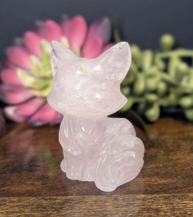May include: A carved rose quartz fox figurine. The fox is light pink and has detailed features. The figurine is sitting on a wooden surface, with a blurred pink flower and green foliage in the background.