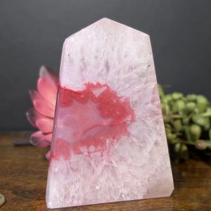 May include: A translucent, obelisk-shaped crystal with a pale pink hue and striking red veining. The polished stone has a flat top and a rectangular base. The crystal is displayed on a wooden surface, with a pink flower and greenery in the background.