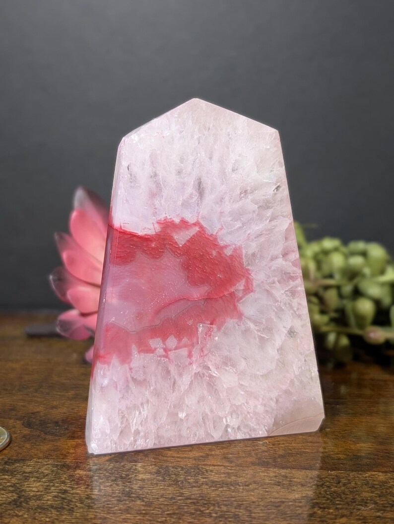 May include: A translucent, obelisk-shaped crystal with a pale pink hue and striking red veining. The polished stone has a flat top and a rectangular base. The crystal is displayed on a wooden surface, with a pink flower and greenery in the background.