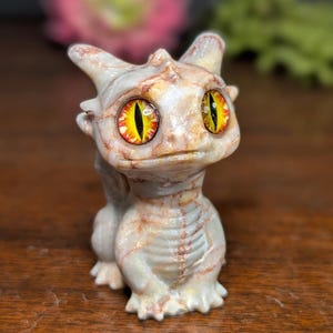 Pode incluir: Pequena figura de drag&atilde;o com um efeito marmoreado bege claro e castanho. O drag&atilde;o tem olhos grandes com &iacute;ris amarelas e laranja. Est&aacute; numa posi&ccedil;&atilde;o sentada, com as asas dobradas e as garras vis&iacute;veis. Objeto decorativo.