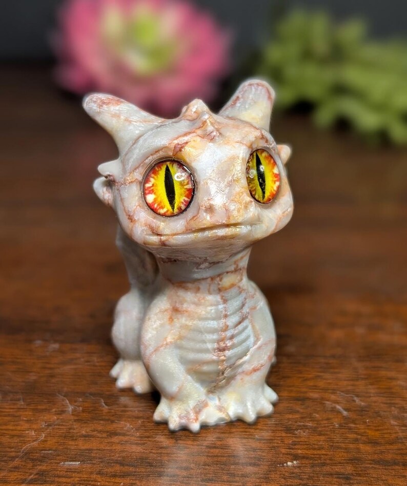 Pode incluir: Pequena figura de drag&atilde;o com um efeito marmoreado bege claro e castanho. O drag&atilde;o tem olhos grandes com &iacute;ris amarelas e laranja. Est&aacute; numa posi&ccedil;&atilde;o sentada, com as asas dobradas e as garras vis&iacute;veis. Objeto decorativo.