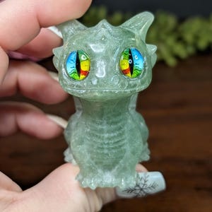 Pode incluir: Uma pequena figura de drag&atilde;o verde com olhos grandes e coloridos. O drag&atilde;o &eacute; esculpido em um material transl&uacute;cido, e seus olhos apresentam um arco-&iacute;ris de cores. A figura &eacute; segurada em uma m&atilde;o, mostrando os detalhes do drag&atilde;o.