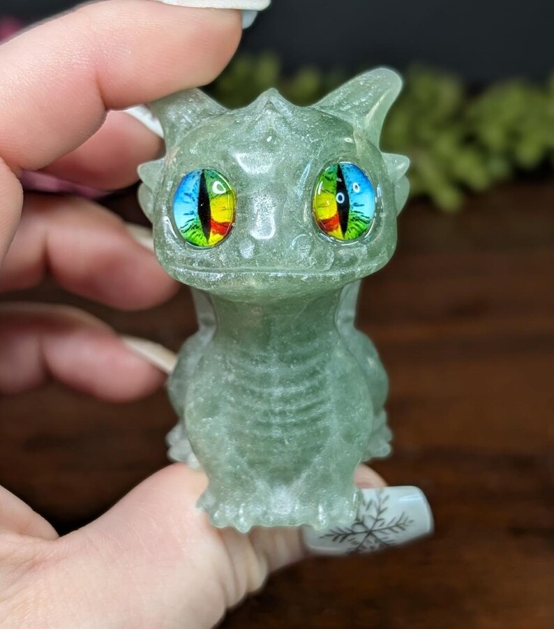 Pode incluir: Uma pequena figura de drag&atilde;o verde com olhos grandes e coloridos. O drag&atilde;o &eacute; esculpido em um material transl&uacute;cido, e seus olhos apresentam um arco-&iacute;ris de cores. A figura &eacute; segurada em uma m&atilde;o, mostrando os detalhes do drag&atilde;o.
