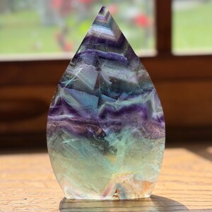 May include: A polished, flame-shaped fluorite crystal carving featuring a gradient of translucent colors, transitioning from deep purple and violet at the pointed top to pale green and clear tones at the flat, stable base with visible internal fractures.