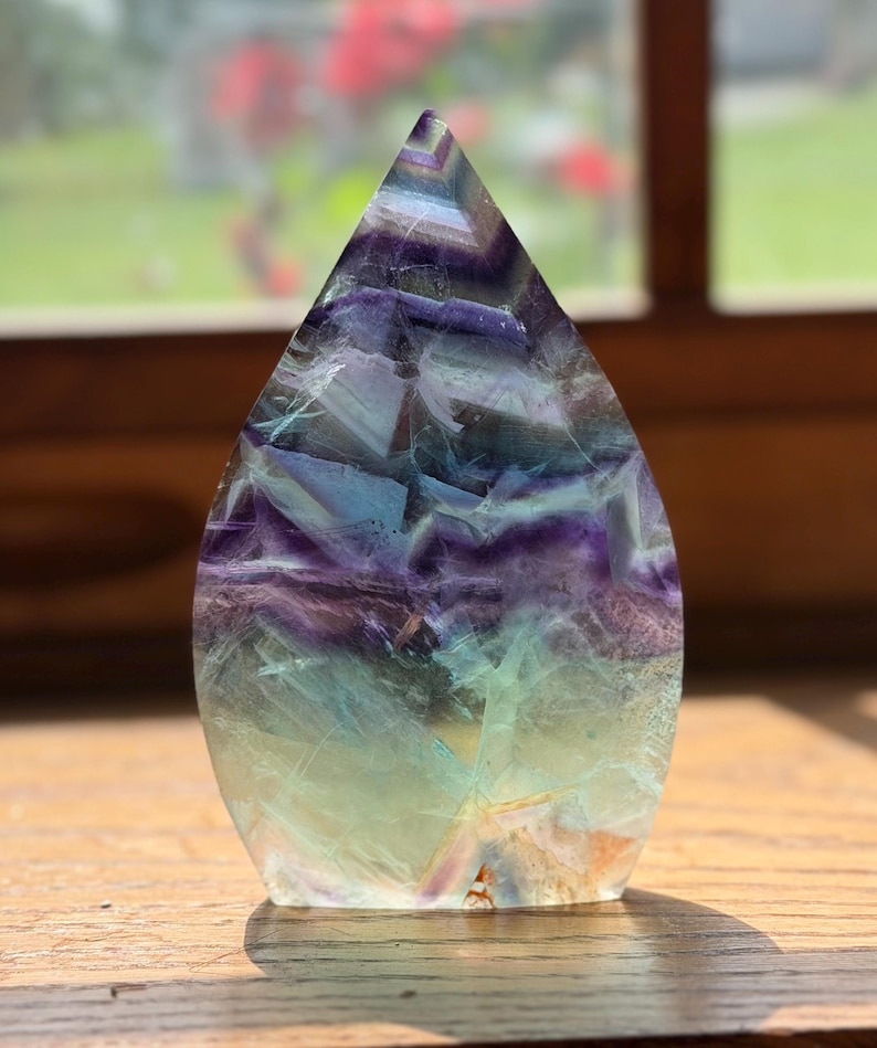 May include: A polished, flame-shaped fluorite crystal carving featuring a gradient of translucent colors, transitioning from deep purple and violet at the pointed top to pale green and clear tones at the flat, stable base with visible internal fractures.