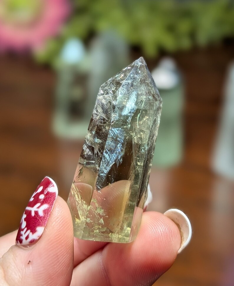 May include: A light green, translucent crystal with a pointed tip. The crystal has internal patterns and inclusions, held between fingers. The background is blurred, with hints of other crystals and greenery.