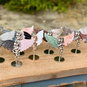 May include: A collection of decorative angel figurines with colorful, patterned wings. Each figurine has a silver-colored body and is mounted on a small, round base. The wings display a variety of colors, including pink, green, and blue.