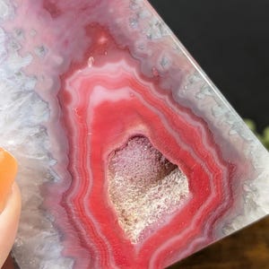 May include: A polished, triangular-shaped agate stone with concentric bands of red, pink, and white. The center features a cavity with a crystalline interior. The stone is held by a person.