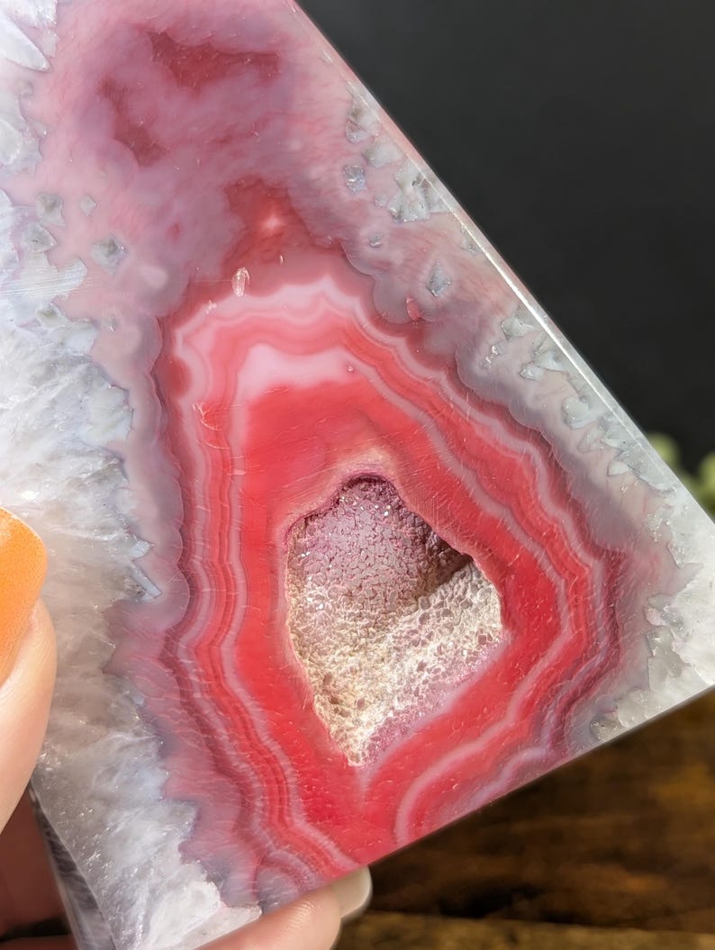 May include: A polished, triangular-shaped agate stone with concentric bands of red, pink, and white. The center features a cavity with a crystalline interior. The stone is held by a person.