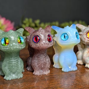 Pode incluir: Uma cole&ccedil;&atilde;o de pequenas figuras de drag&otilde;es detalhadas em v&aacute;rias cores. Os drag&otilde;es t&ecirc;m olhos grandes e coloridos e s&atilde;o feitos de diferentes materiais, incluindo verde, roxo, azul e bege. Est&atilde;o em p&eacute;, com asas e um corpo detalhado.