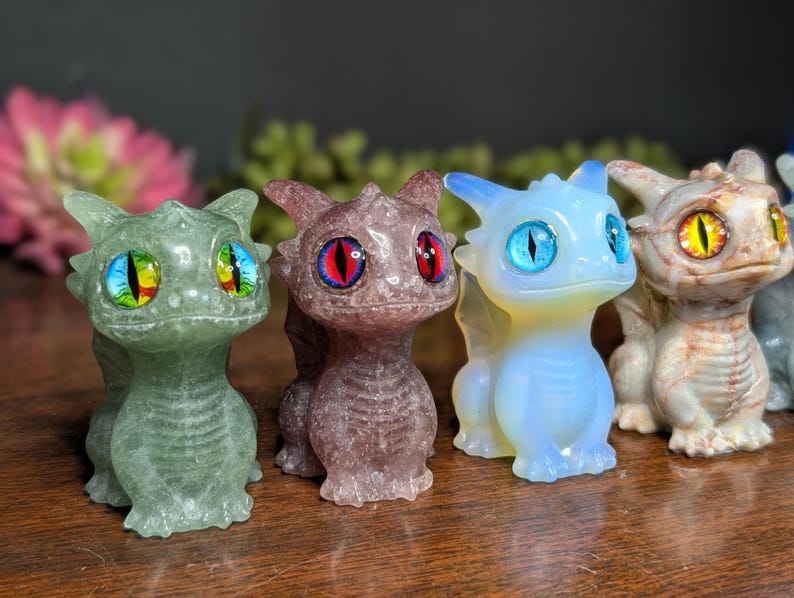 Pode incluir: Uma cole&ccedil;&atilde;o de pequenas figuras de drag&otilde;es detalhadas em v&aacute;rias cores. Os drag&otilde;es t&ecirc;m olhos grandes e coloridos e s&atilde;o feitos de diferentes materiais, incluindo verde, roxo, azul e bege. Est&atilde;o em p&eacute;, com asas e um corpo detalhado.