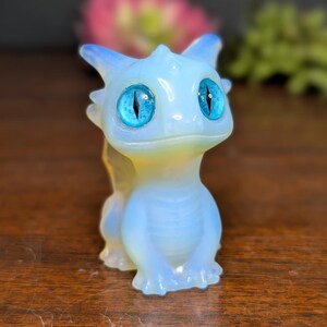 Pode incluir: Pequena estatueta de drag&atilde;o azul claro com grandes olhos azuis brilhantes. O drag&atilde;o tem um acabamento suave e opalescente com sutis destaques amarelos e azuis. A estatueta est&aacute; em posi&ccedil;&atilde;o sentada, com as patas juntas.