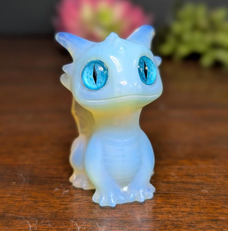 Pode incluir: Pequena estatueta de drag&atilde;o azul claro com grandes olhos azuis brilhantes. O drag&atilde;o tem um acabamento suave e opalescente com sutis destaques amarelos e azuis. A estatueta est&aacute; em posi&ccedil;&atilde;o sentada, com as patas juntas.