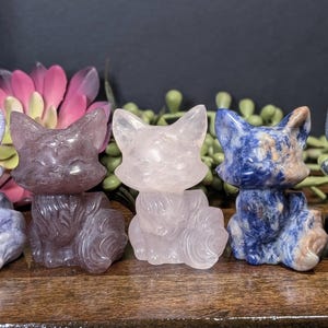 May include: Five carved crystal cat figurines in various colors: blue, purple, pink, blue and white, and gray. Each cat is sitting with its tail curled. The background includes a pink flower and green plants.