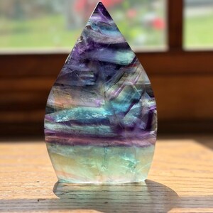 May include: A polished, flame-shaped fluorite crystal sculpture featuring horizontal bands of deep purple, vibrant teal, and pale green. The translucent stone displays natural internal fractures and color variations throughout its smooth surface.