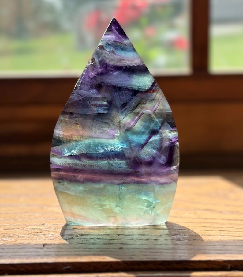May include: A polished, flame-shaped fluorite crystal sculpture featuring horizontal bands of deep purple, vibrant teal, and pale green. The translucent stone displays natural internal fractures and color variations throughout its smooth surface.