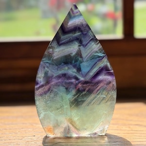 May include: A teardrop-shaped fluorite crystal specimen featuring a polished surface with distinct color banding in shades of deep purple, translucent blue, and pale green, displaying natural internal fractures and geometric chevron patterns.