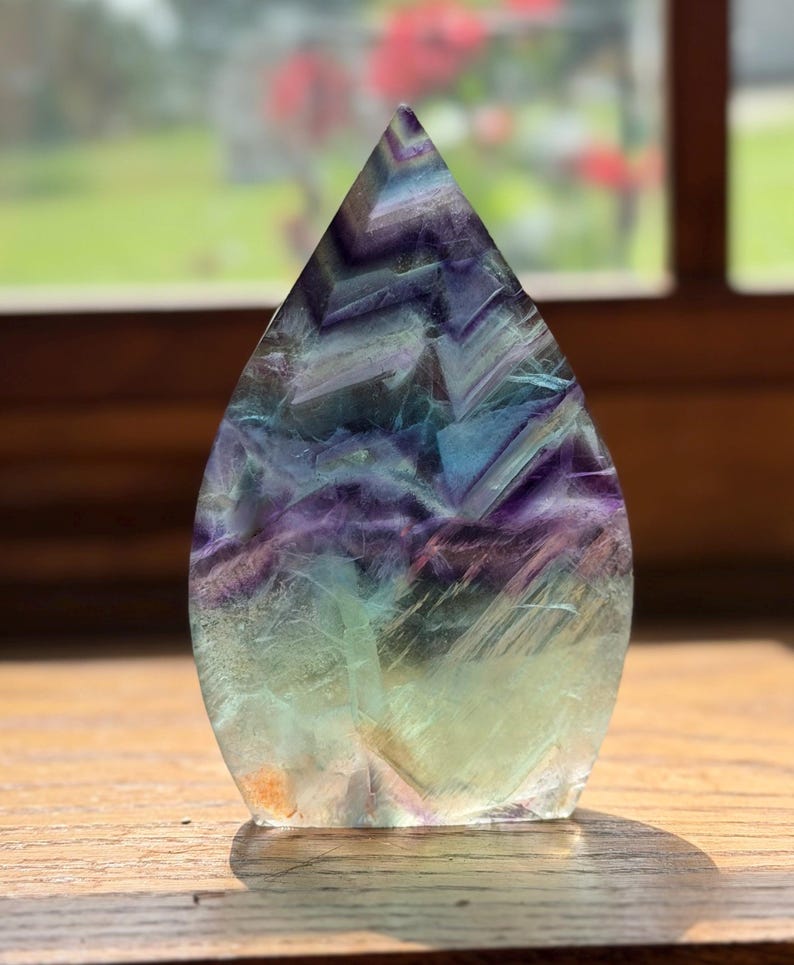 May include: A teardrop-shaped fluorite crystal specimen featuring a polished surface with distinct color banding in shades of deep purple, translucent blue, and pale green, displaying natural internal fractures and geometric chevron patterns.