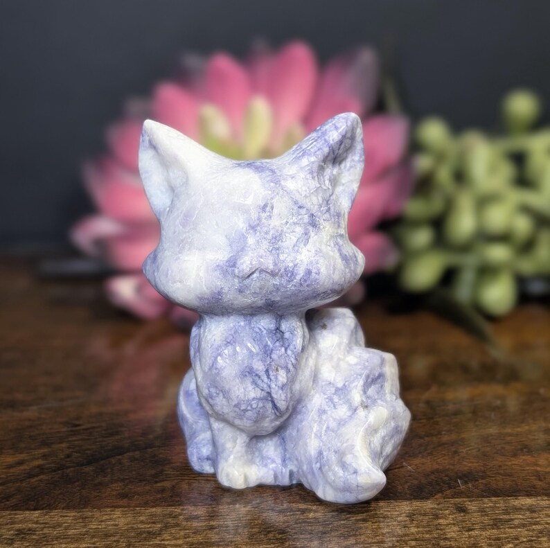 May include: A carved stone cat figurine in shades of blue and white. The cat is sitting upright with its tail curled. The figurine has a smooth, polished finish and is set against a blurred background of pink and green foliage.