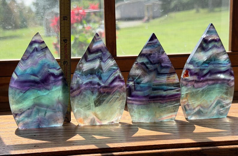 May include: Four flame-shaped fluorite crystals featuring banded patterns of purple, blue, and green. A yellow measuring tape is positioned next to the first crystal, showing a height of approximately 5 inches for the polished stone specimens.