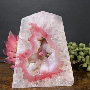 May include: A pink and white geode crystal with a polished, angular shape. The geode features a cavity lined with sparkling crystals and a vibrant pink and white banding pattern. The crystal sits on a wooden surface.