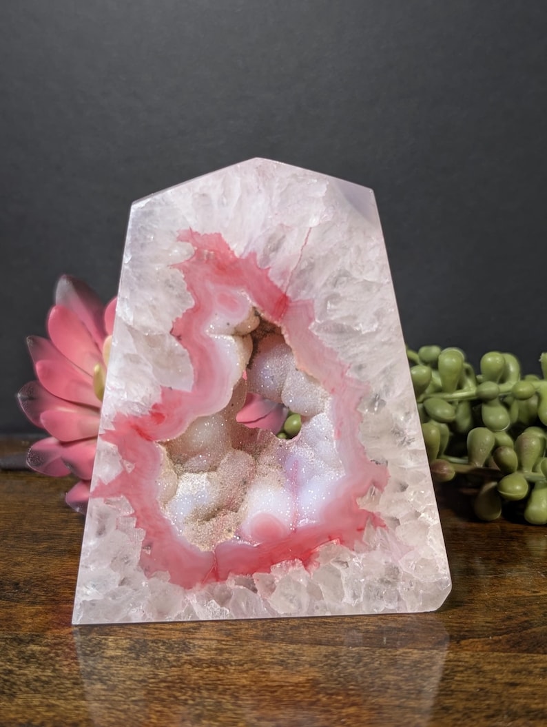 May include: A pink and white geode crystal with a polished, angular shape. The geode features a cavity lined with sparkling crystals and a vibrant pink and white banding pattern. The crystal sits on a wooden surface.