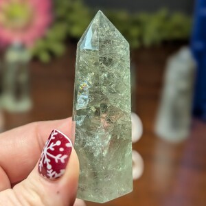 May include: A pale green crystal point with a faceted, pointed top. The crystal is held in a hand with a red and white snowflake design on the fingernail. The crystal has internal fractures and inclusions.